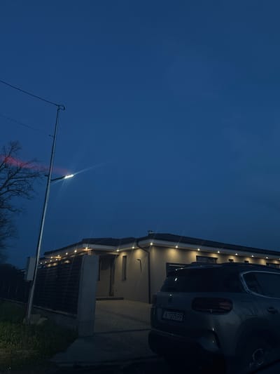 At dusk, the image shows a two-story residential building with outdoor lighting, possibly in Trastikovo, Bulgaria. The building is on the right side of the frame, and a car is parked near its entrance. A street light extends from the left of the frame towards the building. A leafless tree is visible beneath the street light. The sky is dark blue. The license plate on the car is A 1275 PP.
