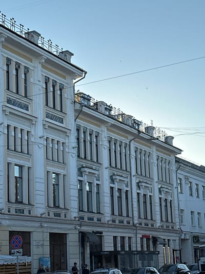 The image captures a side view of a multi-story building with ornate white and light yellow facade, likely located in Yazvitsevo, Russia. The building exhibits architectural details such as columns, decorative window frames with blue tiling, and a wrought-iron railing along the rooftop. A clear blue sky serves as the backdrop, with power lines stretched across the upper portion of the frame. Several small, dark specks are visible in the sky, which could be birds or insects.
In the foreground, a street level view reveals a section of the building's ground floor with windows and doors. A red and blue circular traffic sign with an arrow pointing downwards is mounted on a pole. To the left, a stack of wooden pallets is partially visible. A few people are present on the sidewalk, appearing as dark silhouettes, engaged in unspecified activities. Cars are parked along the street. The overall scene suggests an urban environment during daylight hours, possibly late afternoon or early morning due to the lighting.