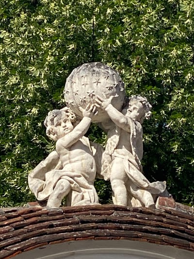 This is a photograph of a stone sculpture of two cherubic figures interacting with a globe. The figures are draped in flowing robes and appear to be straining to hold or manipulate the globe, which is decorated with star-like patterns. The sculpture is positioned outdoors, with lush green foliage serving as the backdrop. A portion of a textured, possibly wooden or tiled, structure is visible on the right side of the image, adding a rustic element to the scene. The lighting suggests it is daytime, with sunlight casting shadows and highlighting the details of the sculpture and the surrounding leaves. The overall impression is one of artistic endeavor and classical representation within a garden setting. There is no visible text in the image.