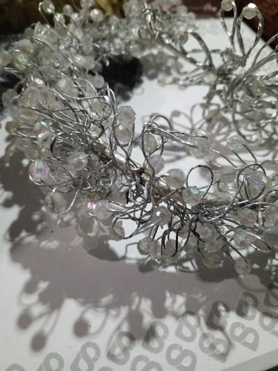A close-up showcases a delicate crown constructed of silver wire adorned with numerous small, iridescent clear beads. The wire is intricately twisted and looped, creating a wiry, spidery effect. The beads cluster densely, reflecting light with subtle rainbow hues. The crown casts a complex shadow on a surface patterned with a repeating "S" motif. The overall impression is one of fragile elegance and handmade artistry.