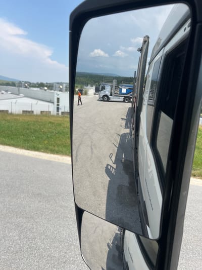 The image is a reflection in a truck's side mirror, showing a scene in Jandelsbrunn, Germany. The mirror is split into two parts, with the larger, primary mirror reflecting a road and a person, and the smaller convex mirror reflecting a wider view of the surrounding area.
In the primary mirror, a person wearing an orange vest is visible standing on an asphalt surface. The person is facing away from the viewer, with their back towards the camera. The reflection also shows parts of a white truck and a trailer, with some metal structures attached to the trailer. Shadows are cast on the asphalt, suggesting it is daytime.
The smaller convex mirror provides a broader perspective, showing more of the road, grassy areas, and buildings. In the distance, several industrial-looking buildings with white facades and dark roofs are visible. Green fields and trees are scattered across the landscape, and a cloudy sky with patches of blue suggests a partially sunny day. The overall setting appears to be an industrial area or a large parking lot. The orientation of the image is such that the top of the mirror is to the left, and the bottom is to the right, implying the photo was taken in a vertical orientation. No discernible text is visible in the reflection.