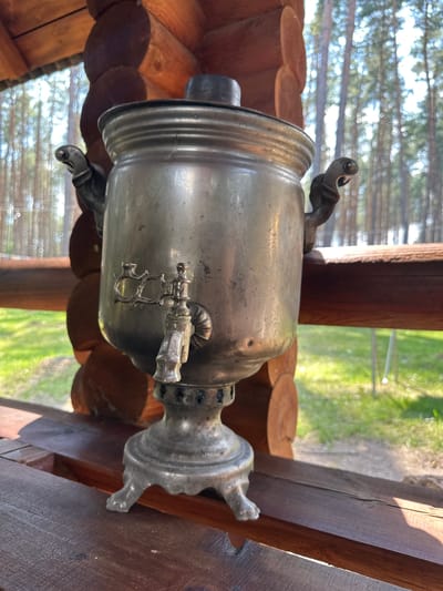 A silver samovar sits on a wooden table outside a log cabin, with a blurred background of trees. The samovar is ornate, with two handles and a spigot. The table appears to be part of a picnic area or outdoor seating space. The overall setting suggests a rural or natural environment, possibly a dacha or a tourist location.