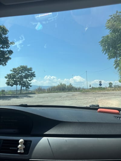 The image captures a static scene from within a vehicle in Marikostinovo, Bulgaria, looking outward through the windshield. The vehicle's dark, textured dashboard occupies the lower portion of the frame. A digital display on the dashboard indicates "(!) Run Flat Indicator" with text detailing a "Run Flat Indicator failure." A small, white, stylized humanoid figurine is affixed to an air vent on the left side of the dashboard, while an orange rectangular object rests on the far right. Reflections of interior elements, including what appears to be a metallic can and other indistinct items, are visible on the windshield glass.
Outside, under a clear blue sky dotted with scattered white cumulus clouds, an unpaved or gravely road surface is visible in the immediate foreground, leading into the mid-ground. Green foliage from trees partially frames the view on the left and right. A prominent, bushy green tree stands centrally to the left of the road. Further right, a utility pole with overhead wires is present, along with more scattered dry vegetation. Two circular regulatory road signs, one predominantly blue with a red diagonal stripe and the other blue and white, are visible in the mid-distance on the right side of the road. In the far background, faint outlines of rolling hills or mountains are discernible beneath the horizon line of more distant vegetation. No people are visible.