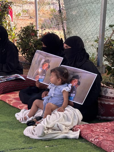 The image depicts an outdoor scene in Carmel, Palestinian Territory. Three individuals, primarily women, are seated on the ground, which is covered with green artificial turf and red and white patterned mats.

In the foreground, two women are seated. Both are dressed in black abayas and niqabs, obscuring their faces. The woman on the right holds a young child, approximately a toddler, who is seated on a white blanket on her lap. The child is wearing a light blue and white striped sleeveless romper dress with a collar and bow, and white sandals, and appears to be resting with eyes closed.

Both women in the foreground are holding identical printed photographs. Each photo displays a smiling young male with dark hair, wearing a dark top with "NIKE KE" printed in light blue. The subject's face and shirt are speckled with what appears to be white debris, and reddish marks are visible on his cheek. Additional printed materials are visible on the mat beside the woman on the left.

The background features a chain-link fence, partially obscured by a green mesh screen or tarp. Sparse vegetation, including bushes with reddish blooms, is visible behind the fence. A light-colored vertical support pole stands near the fence. A red and white flag is partially visible in the far left background.