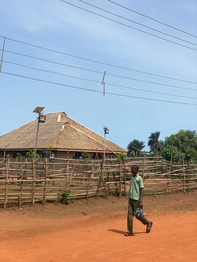 The image depicts a man standing on a reddish-brown dirt path in Ikoga, Nigeria. The man is wearing a light green patterned shirt, dark pants, and sandals, with a bag slung across his body. He is standing parallel to a wooden fence that lines the path, next to a building with a thatched roof. Two streetlights are mounted on poles near the building. In the background, palm trees and other greenery are visible. Power lines run across the clear, blue sky above the scene.