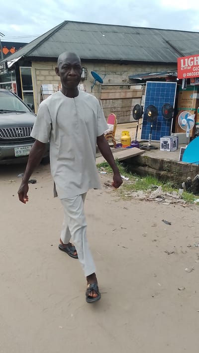 In Agbarho, Nigeria, a bald Black man walks forward in a light-colored agbada and matching trousers. He is wearing black slide sandals. The man is walking on a dirt road next to a dark-colored Lexus SUV. Behind the man and the SUV, there is a building with a corrugated metal roof. In front of the building, there are several items for sale, including solar panels, fans, and a gas cylinder. The sky is overcast.