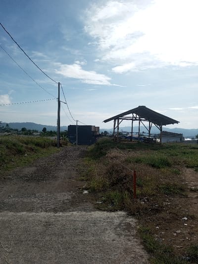 A dirt path leads towards a pavilion under a bright, partly cloudy sky. Adjacent to the path, overgrown grass and weeds blend into the landscape. A dark structure stands near the pavilion, partially hidden by vegetation. Power lines cut across the upper left, extending from a utility pole. Distant hills are visible beneath the sky, completing the outdoor scenery.