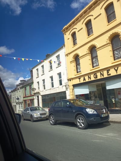 The image shows a street scene in Millstreet, Ireland, on a sunny day with a blue sky and some clouds. On the right side of the street, a yellow building with arched windows and decorative molding is visible. Above the windows, a sign reads "TANGNEY". In front of the building are large windows displaying various items. To the left of the yellow building are several other multi-story buildings, painted in white and green. Some of these buildings have satellite dishes attached to their facades. A string of colorful triangular flags is strung across the street between the buildings.
On the street, there are two cars. A dark blue Nissan Qashqai is in the foreground on the right, and a silver Mercedes-Benz sedan is in the middle ground on the left. The Mercedes-Benz has a license plate that reads "04-C-1640". The image appears to be taken from inside another vehicle, as the left side of the frame shows the interior of a car, including a side mirror and part of the windshield. The street is paved with asphalt, and a white line marks the center of the road. The overall impression is of a typical Irish town street.