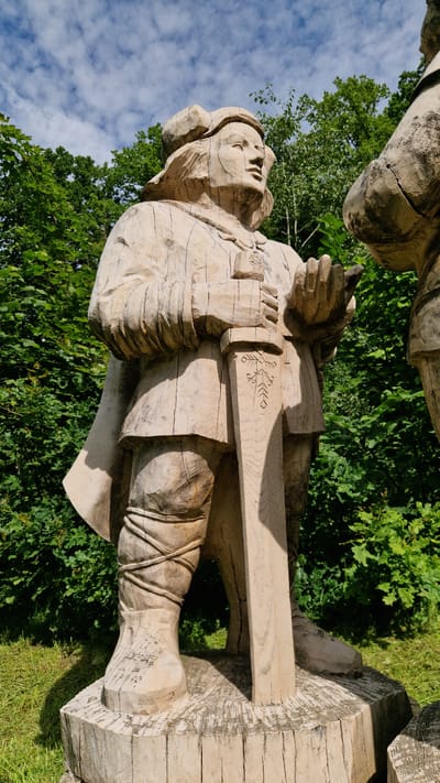 This is a vertical, eye-level, medium shot of a light-colored wooden statue of a man dressed in historical attire, standing in Embūte, Latvia. The statue is carved with visible wood grain and cracks, giving it a rustic appearance. The man is wearing a rounded cap, a cloak over his shoulders, and a tunic. His left hand holds a sword that is planted vertically in front of him. The sword has carved decorative elements on its blade. His right hand is slightly raised and open, as if in conversation or greeting. His gaze is directed upwards and to the right, suggesting he is looking at someone or something outside the frame.

The statue stands on a wooden base, which itself is atop a thick log. Behind the statue, dense green foliage of trees and bushes creates a natural backdrop. The sky above is blue with scattered white clouds. To the right of the main statue, a portion of another similar wooden statue is visible, suggesting they are part of a larger scene or collection. The lighting indicates it is daytime, with sunlight casting shadows on the statue and the ground. The overall impression is one of a historical or folkloric representation displayed outdoors in a natural setting.