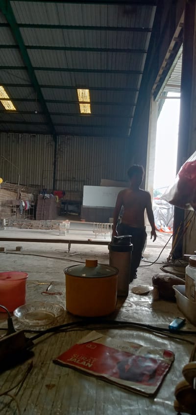 The image depicts an interior scene in a warehouse in Sumput, Indonesia. A shirtless man in dark pants is walking towards the doorway of the warehouse. In the foreground, a cluttered table has a red plastic container, a yellow pot, a steel container, an ashtray, cords, a lighter, and a red and white packet labeled "Surat Jalan." Behind the man, rows of materials are stacked. Clothes hang to dry on a rack near the stacked materials. The warehouse features a corrugated metal roof with visible skylights. The light suggests it is daytime.