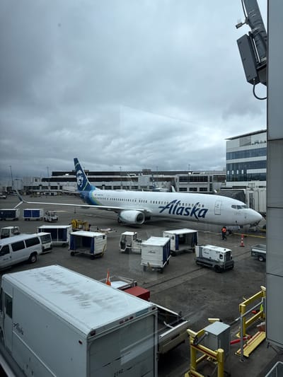 This is a vertical, full-shot photograph taken from an elevated perspective, likely from inside an airport terminal looking out at the tarmac. The image captures an Alaska Airlines airplane parked at a gate, with several pieces of ground support equipment surrounding it.
The main subject is a white Alaska Airlines Boeing 737 aircraft, with its distinctive blue and green logo prominently displayed on the tail and the word "Alaska" written in large blue letters along the fuselage. The plane is connected to a jet bridge, indicating it is either preparing for departure or has recently arrived.
Around the aircraft, various objects and vehicles are visible. These include baggage carts loaded with luggage, a tug vehicle, and a larger white cargo truck. A yellow passenger boarding staircase or platform is also visible, positioned near the front of the aircraft. In the background, an airport terminal building with a modern architectural design featuring large glass windows can be seen.
The scene is set at an airport tarmac, with the aircraft and ground equipment arranged in a typical operational layout. The time of day appears to be overcast, with a gray and cloudy sky dominating the upper portion of the image. The weather seems to be somewhat gloomy or perhaps just a typical cloudy day in SeaTac, United States.
There is visible text on the aircraft, including "Alaska" and "N407AS" on the fuselage and tail. The cargo truck also has "Alaska" printed on its side. Some of the baggage carts also show the "Alaska" logo and identifying numbers.
No specific human activity is clearly discernible, beyond what might be implied by the operational nature of the airport scene. The overall impression is one of a busy, functional airport environment under cloudy skies.