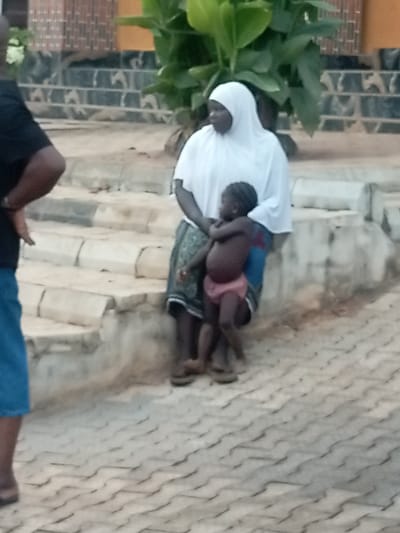A woman wearing a white head covering stands in front of a set of concrete stairs, holding a young child. The child wears a pink diaper. The woman is wearing a patterned skirt, and sandals. A man wearing a dark shirt and blue shorts stands to the left. Behind the woman, there is a large green plant, and a patterned brick building. The ground is paved with grey brick stones.