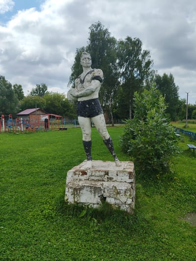 The image shows a weathered statue of a male athlete holding a ball, standing on a crumbling brick pedestal in a grassy park area. The statue is made of concrete and is painted white with black accents on the shorts and leg bands. The athlete has a muscular build and a determined expression. Behind the statue, a playground with red metal structures is visible, along with a building with a red roof. Tall trees with green foliage line the background, and the sky is overcast with gray clouds. The ground is covered in lush green grass with scattered small yellow flowers. The overall setting appears to be a public park or recreational area in Drezna, Russia, likely a sports-themed monument.