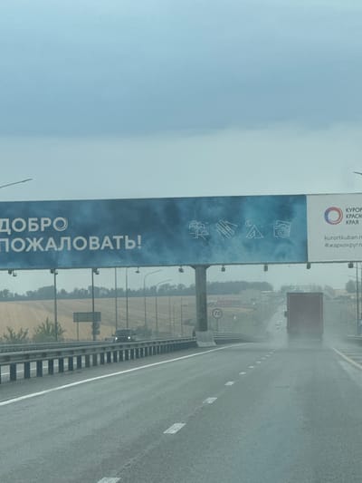 An overcast, hazy sky looms above a highway. A large billboard is visible, displaying the words "ДОБРО ПОЖАЛОВАТЬ!" (Welcome!) in large white letters against a blue, misty background. To the right of this text, the billboard shows several white icons: a palm tree, skis, a tent with a crescent moon and star, and a camera. Further to the right, a circular logo with concentric rings of orange and purple is displayed, followed by text in Russian. Beneath the icons and logo, a website address "kurortkuban.ru" and a hashtag "#жаркокруглый" are printed.
The highway itself is wet, suggesting recent rain, and appears to have multiple lanes. White dashed lines mark the lanes. On the left side of the highway, a metal guardrail runs parallel to the road, with concrete barriers visible in places. Beyond the guardrail, there are trees and agricultural fields, with some structures in the distance. A speed limit sign indicating "70" is visible on the left.
Traffic is present on the highway, with a red truck in the right lane of the distance, creating a spray of water as it drives. A dark-colored car is also visible in the left lane ahead. The overall impression is that of a journey on a highway entering a region, possibly indicated by the welcoming billboard. The location is identified as the city of Tsukerova Balka, Russia.