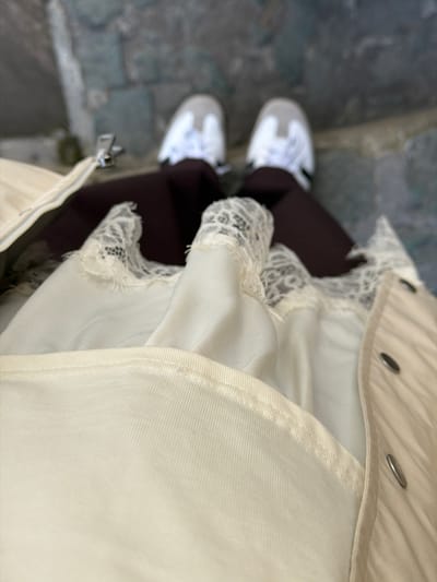An overhead shot depicts a person standing on what appears to be a stone or concrete surface. The person is wearing a beige jacket or coat with visible metal snap buttons, brown leggings, and white sneakers with gray accents. A white dress with lace detailing peeks out from under the jacket. The location is not discernable from the given image.