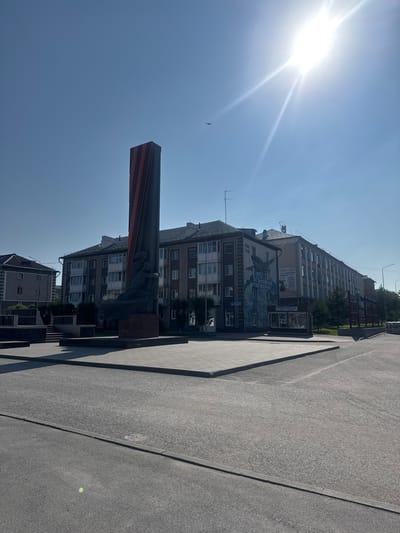 In Belovo, Russia, a monument stands in a public square under a clear blue sky with a bright sun. The monument features a dark-colored, abstract sculpture of a figure embracing a tall, thin obelisk adorned with red stripes. The square is paved with light-colored tiles and bordered by asphalt roads. In the background, several multi-story buildings line the street, some with balconies and windows. One building displays a large mural depicting an airplane and text in Russian, including dates "1941 1945." A small kiosk is visible near the mural. The environment is urban, with a few scattered trees and a street lamp visible. A bird is seen flying in the distance.