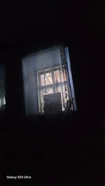 A dark, indoor scene focuses on a window framed by lace curtains. Light from outside the window illuminates a paneled glass window with what appears to be a wooden frame. Through the glass, faint outlines of external structures are visible, possibly a roof or sky. A framed object, possibly a picture or book, sits on the windowsill. The bottom right corner of the image displays the text "Galaxy S23 Ultra," indicating the device used for capture. The overall impression is one of a dimly lit room in Pîrîta, Moldova, with the window as the central focal point.
