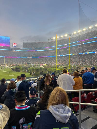 A daytime shot of a packed stadium with many spectators in the stands. The field is visible with yellow goalposts. Some people are in the foreground in stadium seating. A large Pepsi sign is on a screen. Overhead lighting is on.