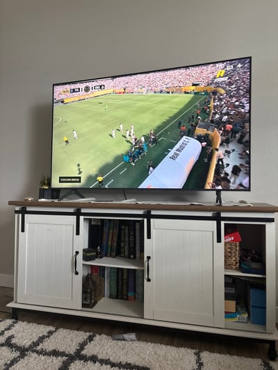 This image depicts an indoor scene, consistent with a residential living area, located in Tigard, United States. A large, flat-screen television dominates the center, displaying a live soccer match. The on-screen score indicates PSG leading Real Madrid (RMA) 3-0 at the 76:50 mark, with a "COOLING BREAK" notification visible in the lower left corner. The broadcast shows multiple soccer players on a green field, a dugout labeled "Real Madrid C.F.", and a dense crowd in the stadium stands. The television rests on a white media console with a dark brown wooden top and black, barn-door-style sliding hardware. The console's open shelving on the viewer's left contains several books, a small dark box, and a golden decorative lantern. The open shelving on the right holds a woven basket containing a red item, footwear (possibly slippers) on the shelf below it, and other stacked items on the bottom shelf. A small green potted plant and a small golden object are positioned on the console's top surface to the left of the television. The floor is covered by a white, textured rug with a gray geometric pattern. A light gray slipper is visible on the rug in the foreground. The wall behind the console and television is plain and light-colored.