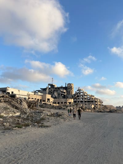 In Jabalia, Palestinian Territories, a broad, sandy area leads towards a heavily damaged, multi-story building. The building, appearing to be a former hotel or large apartment complex, is in a state of severe destruction, with large sections of its structure collapsed, exposing rebar and concrete. Debris, including rubble and twisted metal, is scattered around the building and in the foreground. The sky above is blue with scattered white clouds, suggesting it is daytime. Three young males are walking away from the camera, towards the damaged structure and along the sandy path. The environment is barren with sparse vegetation growing amongst the rubble.
