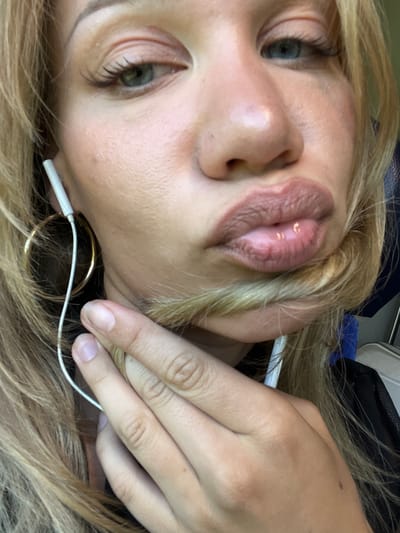 A close-up, head-and-shoulders selfie of a young blonde woman, taken from a low angle. She has green eyes and is wearing large gold hoop earrings and white earbuds. Her lips are slightly parted, and she is holding a lock of her hair with her right hand, obscuring her mouth. The background is blurred and dark, with a hint of blue fabric. There is no indication of the location in Lengdorf, Germany.