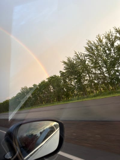 The image captures a view from inside a vehicle, looking towards the side mirror and out the window. A faint rainbow arcs across the pale sky on the left side of the frame. A line of trees, appearing dense and green, runs parallel to a dark asphalt road. The road has white lane markings and appears to be wet, possibly from recent rain. Reflections of the sky and trees are visible in the side mirror, which is black and mounted on the vehicle. The overall impression is a travel scene, likely on a highway, with the natural phenomenon of a rainbow present. The location is identified as Таймурзино, Russia.