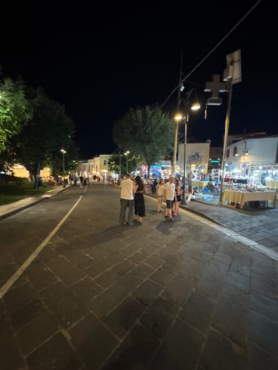 The image depicts a street scene at night in Orosei, Italy. A paved street with cobblestone-like sections and a white line marker runs through the center of the frame. On the right side of the street, there are several market stalls displaying goods, illuminated by overhead lights. People are browsing and interacting at these stalls. On the left side of the street, a grassy area with trees is visible, also illuminated by streetlights. Several people are walking along the sidewalk and street. In the foreground, two individuals stand facing away from the camera, with one person wearing a white top and striped pants, and the other wearing a dark dress. The overall atmosphere suggests a lively evening in a town square or main street with shops and activities.