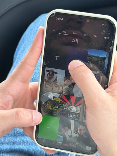 A person holds a cracked-screened iPhone displaying a photo gallery or social media feed with the text "Activity All Favorites" at the top. The phone's screen shows a time of 09:54 and indicators for cellular data ("66"), location ("NEAR"), and categories labeled "MY," "NEW," "TOP," "CORE," and "PPL." The screen also displays a "+" symbol, implying an add or upload function, and the number "3264." Thumb obscures part of the screen. The person's hands are visible, with a denim-like material in the background. The location in the city of Mrgvali Chala, Georgia, is not determinable from the provided image.