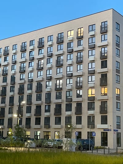 The image shows a modern, multi-story apartment building with a light-colored brick facade. The building has numerous windows and small balconies, many of which have air conditioning units installed below them. Several windows on the upper floors are illuminated with a warm, yellow light, suggesting interior lights are on. The building appears to be located in a developed urban area, with cars parked in front of it, some of which are visible in the foreground. There are streetlights and a security camera mounted on the building. Signs with the letter "P" are visible, indicating parking. The sky above is a clear, pale blue. The lower part of the image shows lush green grass and some trees, suggesting a landscaping element. This scene is located in Ильинское-Усово, Russia.