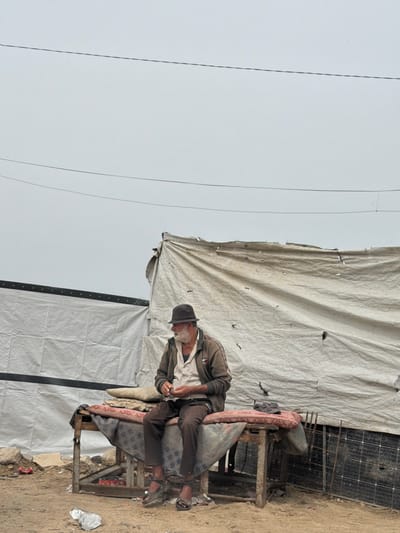 An elderly man sits on a makeshift chair near a shelter constructed from tarps in an outdoor environment. The location is غزة, Unknown. The man wears dark pants, a light shirt, a jacket, and a dark hat. The chair appears to be composed of scavenged materials. A solar panel is present near the shelter. Overhead power lines are visible against a gray sky. Trash is scattered on the ground.