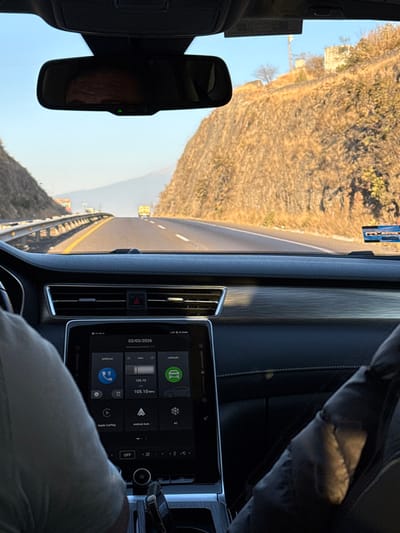 The image captures a first-person perspective from the interior of a vehicle traveling on a highway in San Bernardino Chalchihuapan, Mexico.
The foreground displays the vehicle's dashboard, featuring a large central infotainment screen. The screen indicates the date "03/03/2026," an exterior temperature of "7°C," and active radio tuned to "105.10 MHz." Options for "telefono" (phone), "radio," and "vehiculo" (vehicle), alongside Apple CarPlay, Android Auto, and climate controls, are visible on the interface. Above the screen, a pair of air vents are present. A rearview mirror is mounted centrally at the top of the windshield, reflecting part of a human head and displaying a small green indicator light. On the lower-left, a driver's arm and shoulder, clad in a light-colored shirt, are visible. On the lower-right, a passenger's arm, dressed in dark, possibly padded clothing, is partially visible. An "MX pass" toll transponder sticker is affixed to the upper-right portion of the windshield.
The exterior environment depicts a multi-lane highway stretching into the distance. A metal guardrail parallels the left side of the road. Both sides of the highway are bordered by steep, rocky hills or mountains covered with sparse, dry vegetation, including some cacti. A yellow bus or large truck is visible in the middle distance on the highway. On the right hillside, a telecommunication tower or similar structure and remnants of a stone structure are discernible. The sky is clear and light blue, indicating daylight conditions. The vehicle appears to be in motion.