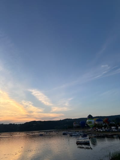 The image depicts a serene sunset over the Jezioro Rożnowskie lake in Gródek nad Dunajcem, Poland. The sky is a gradient of soft blues and oranges, with delicate wispy clouds reflecting on the calm water. Several hot air balloons are visible in the distance, adding a splash of color and a festive atmosphere to the scene. The shoreline is lined with trees and hills, and a collection of boats and small structures suggest a populated area. There are no distinct individuals or interactions captured in the frame, only the quiet beauty of a lakeside evening.