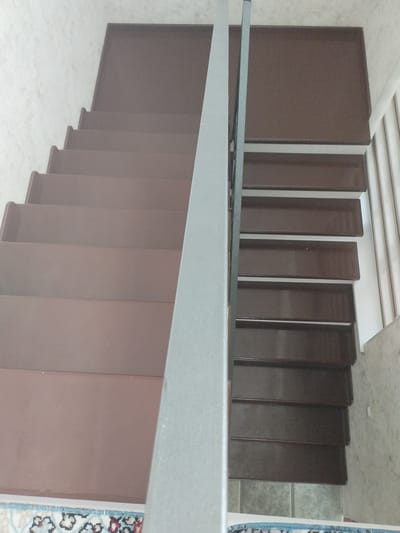 An indoor staircase with brown treads and a silver handrail is shown in the city of Октябрь, Kyrgyzstan. The staircase is L-shaped, with multiple steps visible on both sides of the handrail. The walls are a light, textured beige. A portion of a patterned rug is visible at the bottom of the image.