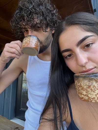 Two individuals are depicted outdoors in Otego, United States. In the foreground, a female with long dark hair and prominent eyebrows holds a speckled ceramic, handleless mug to her mouth, looking directly forward. To her left and slightly behind, a male with dark, curly hair and a beard, wearing a white tank top, holds an identical speckled ceramic mug to his mouth, partially obscuring his lower face. Both subjects appear to be consuming a beverage. The immediate environment includes wooden deck boards at the lower left and a dark-colored building exterior with a reflective window or door panel behind the male. A brown wooden ceiling or overhang is visible in the upper background.