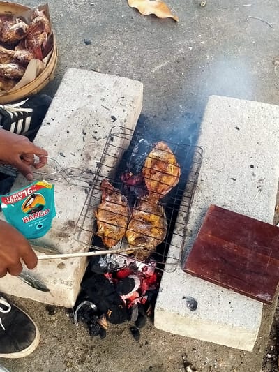 An outdoor grilling scene is captured in Celep, Indonesia, on a concrete or paved ground surface. A makeshift grill is constructed from two light-colored concrete blocks supporting a metal grate. Three whole fish, appearing golden-brown and cooked, are positioned on the grate directly over red-hot charcoal and ash, from which light smoke emanates. To the upper left, a woven bamboo basket holds several pieces of fried fish, partially wrapped in light-colored paper. Two human hands are visible: one hand on the left holds a pair of metal tongs along with a teal-colored pouch labeled "BANGO KECAP MANIS" (sweet soy sauce); the other hand, in the lower-left foreground, holds a thin wooden stick, extending towards the glowing coals. A rectangular, dark brown wooden board rests on the right concrete block. Footwear belonging to two individuals is partially visible: a black shoe with white stripes on the upper left, and a black shoe with white laces on the lower left. A single dry leaf is present on the ground in the upper-middle background.