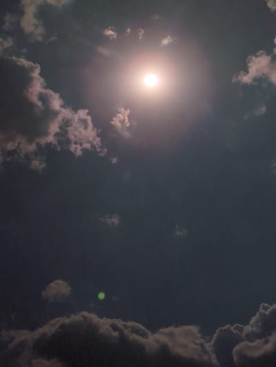 The image depicts a cloudy sky with the sun partially obscured by clouds, creating a bright, hazy halo effect. There are scattered cumulus clouds of varying sizes and densities, some appearing light and wispy, while others are darker and more substantial, particularly on the right side of the frame. The overall impression is of a daytime scene, likely late afternoon or early evening given the lighting and the soft, diffused sunlight. A small, bright green orb, possibly a lens flare or an artifact, is visible in the upper right quadrant of the image. The setting appears to be an outdoor environment, given the focus on the sky. No people, objects, or text are clearly discernible. The colors are muted, dominated by shades of gray, white, and a soft pinkish hue emanating from the sun. The location context provided is Borovan, Bulgaria.