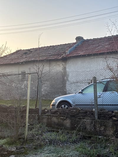 The scene depicts an outdoor residential or rural area in Varbak, Bulgaria, under a pale, clear sky indicative of low-angle sunlight. In the background, a building with a light-grey, textured facade and a weathered, red terracotta tile roof is visible. The roof exhibits areas of disrepair and surface accumulation. A dark grey chimney structure projects from the roofline towards the right. A single, dark, rectangular window is present on the upper left portion of the building's facade. Overhead power lines span the upper section of the image.
A chain-link fence, supported by various posts, runs horizontally across the midground. Behind this fence, a silver or light-grey passenger vehicle, consistent with a station wagon or hatchback, is parked, oriented left-to-right. The vehicle's front passenger side, including the wheel, partial hood, and a frosted windshield and windows, is visible. Sparse, leafless deciduous trees and shrubs are distributed across the midground, both behind and in front of the fence. A slender, bare tree stands centrally behind the fence.
In the lower midground, a low, deteriorating stone wall extends horizontally. The foreground consists of ground covered with sparse, frost-dusted vegetation, primarily short grass and small plants. Additional stones and debris are scattered on the ground near the base of the stone wall. No individuals or discernible activities are present within the frame.