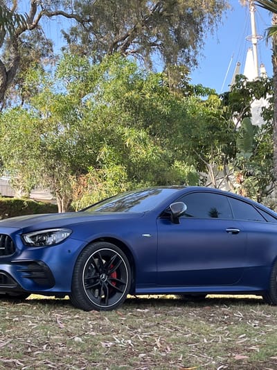 A matte blue Mercedes-Benz C63 AMG, likely a recent model based on design features, is parked on a grassy area. The car faces left, revealing its front and driver's side. Distinct features include black alloy wheels with red brake calipers, tinted windows, and the AMG badge on the front fender. The environment suggests a residential or park setting with trees and vegetation in the background. A boat mast is visible behind the car, indicating proximity to a marina or coastal area. Given the prompt's reference to Germasogeia, Cyprus, it's plausible the scene is set near the Germasogeia dam or the coastal region of Germasogeia. No people are visible.