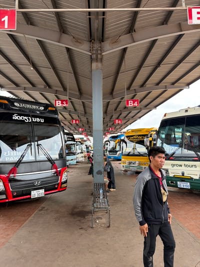 The image depicts a bus station in Phakhao, Laos. In the foreground, a man stands looking to the right. He is wearing a dark grey and black zip-up jacket over a red shirt, and dark pants. Behind him, several buses are parked under a covered shelter. The bus on the left is black with "LUXURY" branding and text in a script. The next bus is blue, followed by a yellow bus with "VIP" and "Mousin" branding. Further back, a white and green bus with "grow tours" written on it is visible. Red signs indicating bus bay numbers (B3, B4, B5, B6, B7, B9, B10, B11, B12, B13) are suspended from the shelter. The ground is paved with concrete and some reddish tiles. The environment appears to be outdoors, with a cloudy sky visible above the shelter. Several other individuals are seated or standing in the background near the buses. The overall scene captures a moment at a busy transportation hub.