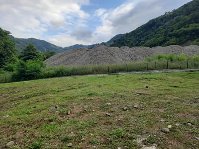 The image shows a vast expanse of raw materials, likely gravel or crushed stone, piled high in a landscape that appears to be near Ayvacık, Turkey. The piles of material are grey and textured, forming irregular mounds that stretch across the middle ground of the image. They are bordered by a green, bushy vegetation line behind which a fence is visible. In the background, lush, green, forested mountains rise steeply, with a partly cloudy sky overhead. The foreground consists of a grassy field, with scattered rocks and sparse patches of bare earth. The scene suggests a quarry, construction site, or a similar industrial or storage area integrated into a natural, mountainous environment. There are no people or vehicles visible in the image.