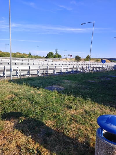 The image depicts a roadside scene on a clear, sunny day. In the foreground, a patch of dry and green grass is visible, with a concrete and blue trash receptacle on the right. Behind the grass is a metal guardrail system, characteristic of a highway or major road. Beyond the guardrail, a multi-lane road stretches into the distance, with streetlights lining the median. A triangular warning sign is posted on the left side of the road, and a circular blue sign with a white arrow indicating "forward" is visible on the right. The background features a tree line and a bright blue sky with scattered white clouds. The presence of highway infrastructure, such as guardrails and traffic signs, suggests a location along a major transport route. Given the context of Sesto Fiorentino, Italy, this scene is likely situated along one of its arterial roads or highways, possibly near an entrance or exit ramp. There are no people or vehicles visible in the image.