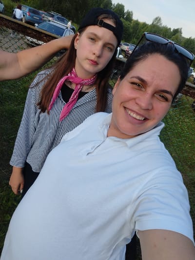 Two individuals, a young female and an adult female, are photographed outdoors in an area resembling a parking lot or gathering space. The young female, positioned behind and to the left of the adult female, has long brown hair, is wearing a black baseball cap, a black shirt, and a vertically striped long-sleeved shirt with a pink bandana tied around her neck. The adult female is in the foreground, facing the camera, and smiling. She has dark hair pulled back, is wearing a white polo shirt, and has sunglasses on top of her head. In the background, several vehicles are visible, and beyond that, a wooded area with trees. A chain-link fence is also present in the scene. The overall lighting suggests an outdoor setting, likely during the day.