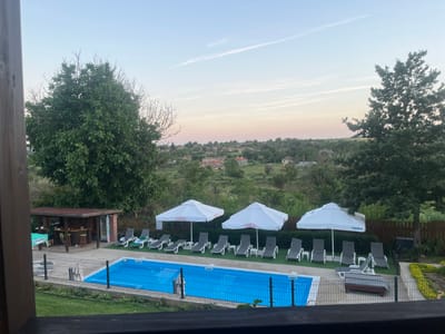 The image shows an outdoor swimming pool area at dusk in General Kiselovo, Bulgaria. The pool is rectangular and filled with bright blue water, surrounded by a black metal fence. Several lounge chairs and white umbrellas with red Coca-Cola branding are neatly arranged on the artificial turf next to the pool. To the left, a wooden bar with a red roof is visible, with a person standing behind it and stools at the counter. In the background, a village with scattered houses and trees is nestled in a gently rolling landscape under a pale sky with wispy clouds. Tall trees frame the scene on the left and right, adding to the natural setting.