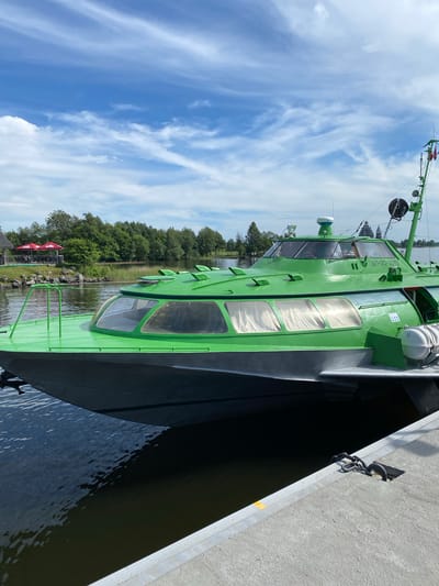 A bright green hydrofoil boat is docked at a concrete pier on a body of water. The boat has a sleek, modern design with large, tinted windows. The hull of the boat is dark gray. The water is calm and reflects the blue sky and white clouds. In the background, lush green trees line the shore. A building with red umbrellas is visible on the left side of the image. The sky is a vibrant blue with scattered white clouds. The image captures a serene daytime scene in Yamka, Russia.