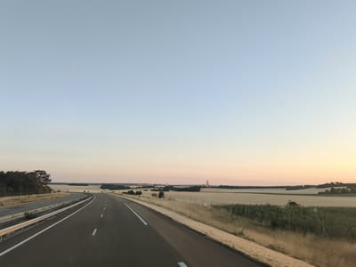The image depicts a highway view in the countryside, possibly near Nitry, France. The perspective is from a vehicle traveling on a paved road with clear white lane markings. On the left, there is a metal guardrail and a grassy embankment with trees. The landscape to the right of the highway features agricultural fields, some appearing to be harvested grain, and a patch of green vegetation, possibly sunflowers. In the distance, there are several wind turbines and structures resembling industrial buildings or towers. The sky is a clear gradient from pale blue at the top to a soft pinkish-orange hue near the horizon, indicating it is either early morning or late evening. The overall scene is serene and characteristic of rural French landscapes.