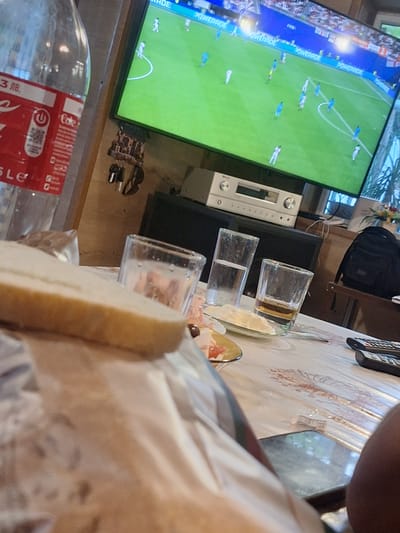 The image shows a person's arm and shoulder in the foreground, obscuring a portion of a table setting. On the table, there are two clear glasses, one partially filled with a light-colored liquid, likely a beverage. A small plate with some food, possibly cheese and fruit, is also visible. A piece of bread or cake lies in front of the glasses.
In the background, a large television screen displays a football match. The game appears to be in progress, with players in blue and white uniforms on a green field. The television is placed on a stand, next to an audio receiver. A black backpack is visible on a shelf above the audio equipment.
The scene suggests a casual gathering or a moment of relaxation, possibly watching a sports event. The lighting indicates it might be daytime or evening. The presence of food and drinks suggests a meal or snack is being enjoyed. There are no clear weather cues.
Visible text includes "POWERADE" and "FIFA" on the television screen. On the bottle in the lower left corner, text appears to be in Cyrillic script and "Coca-Cola" is partially visible. The location context provided is Mihailovo, Bulgaria.