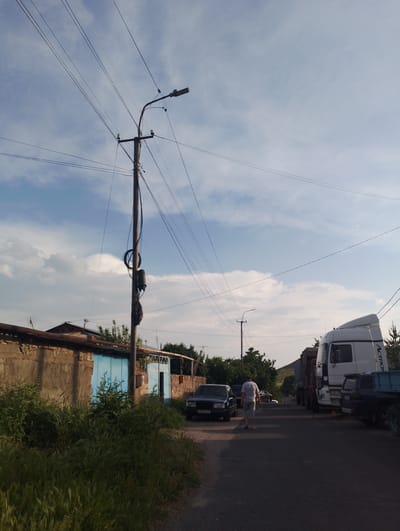 The image depicts a street scene in Nor Geghi, Armenia. A person wearing a white t-shirt and light-colored shorts is walking away from the camera on the right side of the road. Multiple vehicles are present: a dark-colored sedan is parked on the left, a white semi-truck with "Actros" branding is parked on the right, and a blue utility vehicle is parked behind the semi-truck. A smaller, dark-colored vehicle is visible further down the road. A utility pole with numerous wires is centrally located in the frame, with a street light attached. The background features a low stone wall, a building with blue doors, and trees. The sky is partly cloudy, with a blue sky visible. The overall environment appears to be a rural or suburban street.
