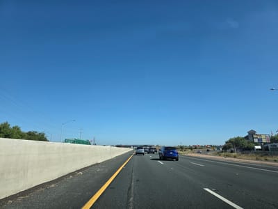On a clear, sunny day in Elk Grove, California, USA, a blue SUV travels down a multi-lane highway. The vehicle is in the right-hand lane, moving alongside other cars. To the left of the highway is a concrete barrier, and beyond that, trees and power lines. On the right side of the highway, there are businesses and signs, including a 76 gas station. The sky is a vibrant blue with no clouds.