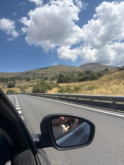 The image captures a view from the passenger side of a car traveling on a road that curves to the left. The road has white dashed lines on the left and a metal guardrail on the right. In the distance, there are rolling hills covered with dry grass and scattered green trees, indicative of a Mediterranean landscape. A vast blue sky with fluffy white clouds fills the upper portion of the frame. A side-view mirror reflects the upper torso of a person wearing a light-colored shirt, with their head tilted slightly downward. The setting appears to be a sunny day in a rural area, possibly near El Espinar, Spain, given the natural terrain and vegetation.