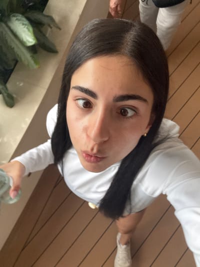 A high-angle photograph captures a young female looking directly upwards at the camera, exhibiting a cross-eyed gaze and pursed lips. She has long, dark hair with a visible part, dark eyebrows, and brown eyes. A small gold stud earring is present in her right ear. She is dressed in a white long-sleeved shirt and a light-colored shoe is visible on her right foot. Her left hand, with light-colored fingernail polish, holds a clear plastic water bottle. The immediate environment consists of a light brown, horizontally planked floor, consistent with a deck surface. In the upper left background, green foliage is partially visible adjacent to a light-colored vertical structure. The upper right background shows the lower legs and feet of a second individual, wearing white pants and light-colored footwear. A dark object, possibly a bag, rests near the second individual's leg. The scene is located in the city of Berry Islands, Bahamas.